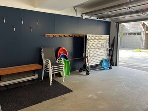 A garage with a bench, a folding chair, a rack with helmets, and a cart with a blue container.