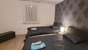 Een slaapkamer met twee bedden, een bankje, een raam met dunne gordijnen en een behang met patroon.