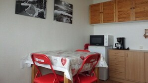 A small kitchen with a microwave, a coffee maker, and wooden cabinets. There is a table with a heart-patterned tablecloth and red chairs. Two black and white photographs are on the wall. - Studio at the foot of the slopes, for 2 people. (Villarembert)