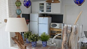 Une cuisine avec des meubles blancs, un four à micro-ondes et un réfrigérateur. On trouve sur le plan de travail des objets décoratifs, notamment une lampe, des plantes en pot et un vase en verre contenant des morceaux de bois flotté.