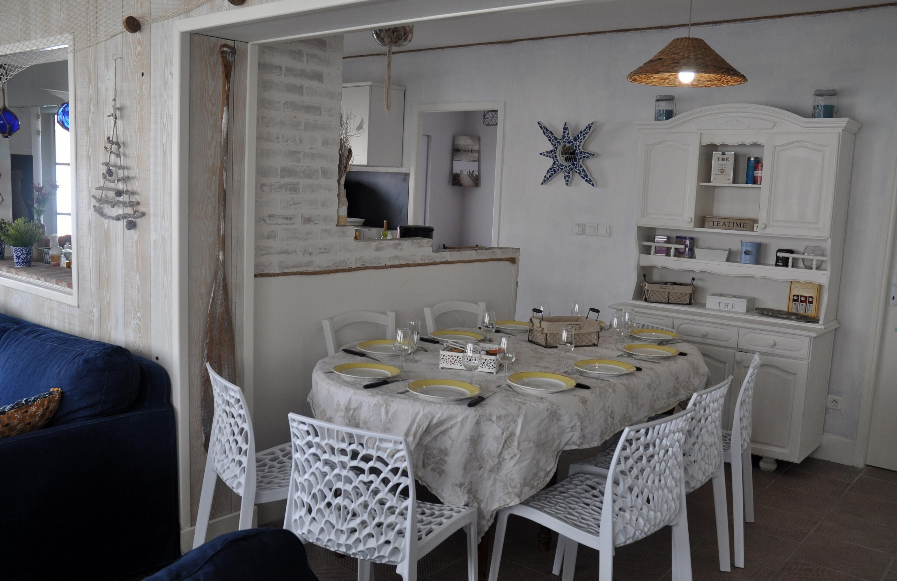 A dining area with a round table set for six, surrounded by white chairs. A white cabinet with shelves is visible in the background.