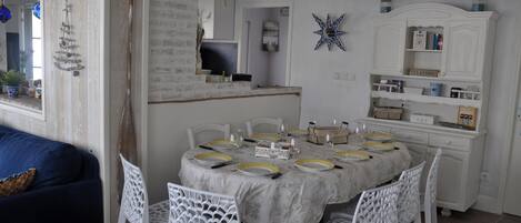 A dining area with a round table set for six, surrounded by white chairs. A white cabinet with shelves is visible in the background.