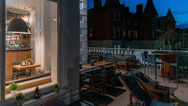 A rooftop bar with wooden tables and chairs, a bar area with a hanging light, and a view of a historic building with pointed roofs. - The Strand Hotel, the former residence of Oscar Wilde (Bray)