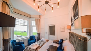 A hotel room with a large bed, two blue armchairs, a television, a window with a view of a green field, and a modern chandelier. - The Strand Hotel, the former residence of Oscar Wilde (Bray)