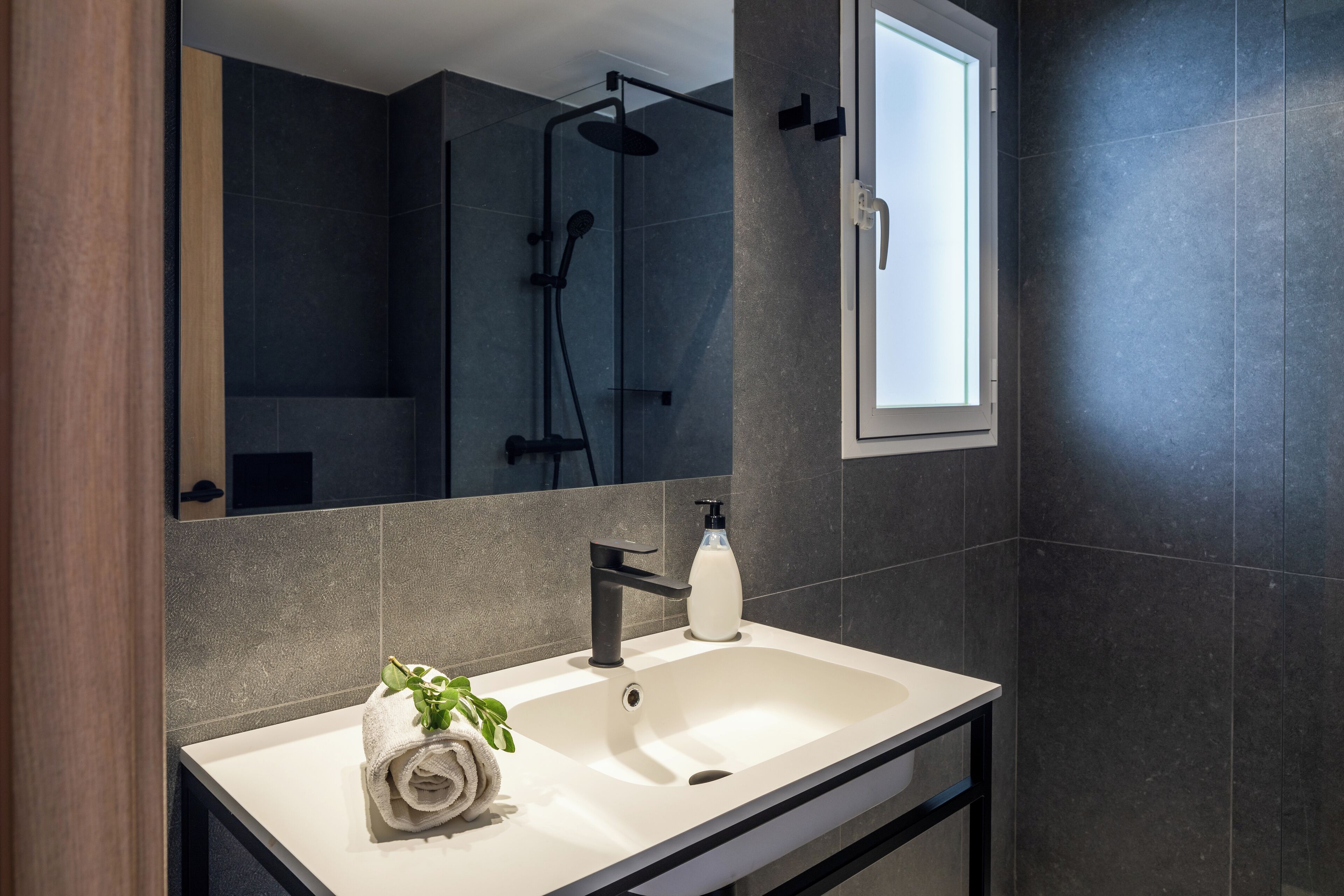 Un baño moderno con un espejo grande, un lavamanos blanco, un grifo negro, un cabezal de ducha de mano y una ventana.