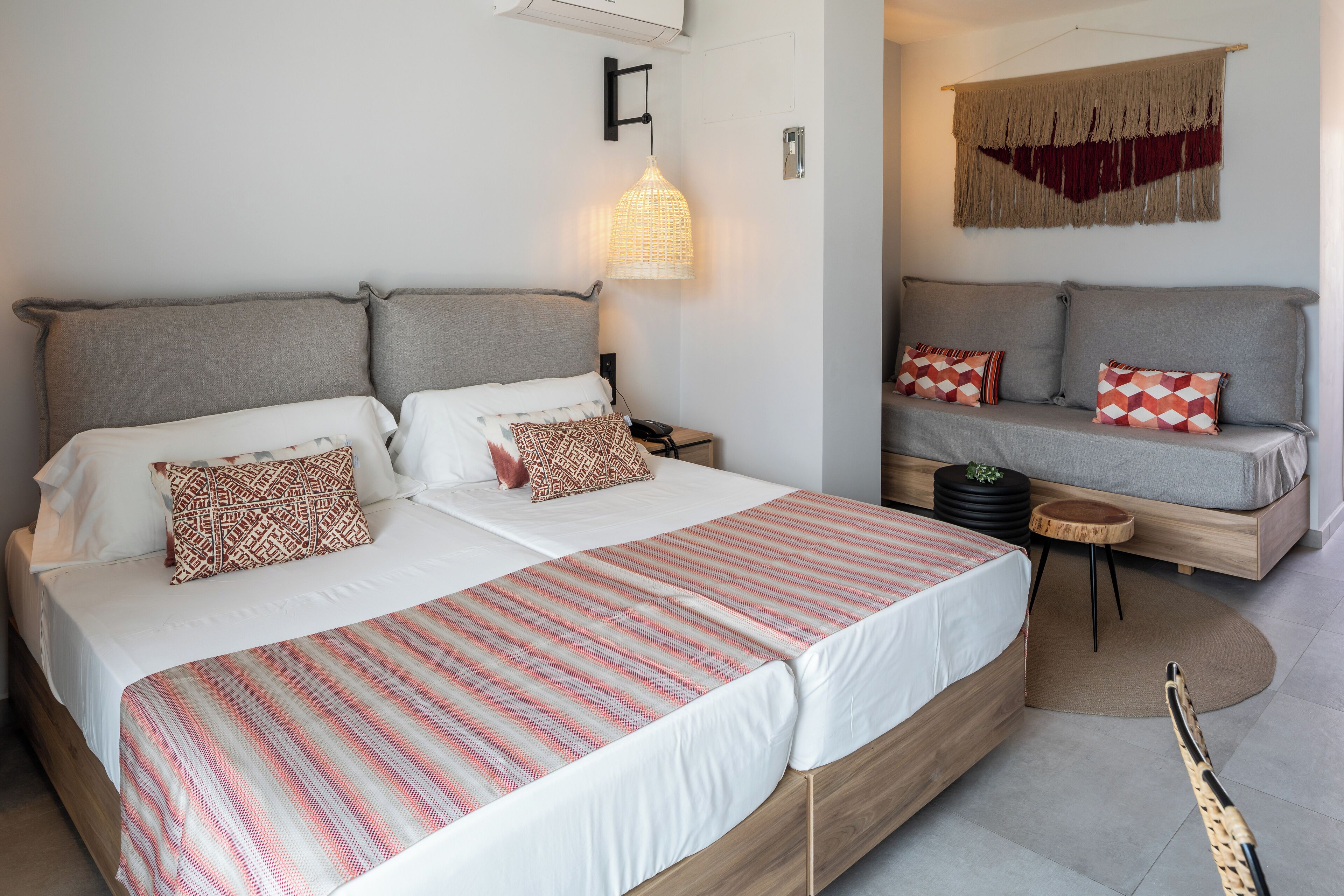 A double bed with a striped bedspread, two bedside tables, a wall-mounted light, a sofa with patterned cushions, and a small round table with a plant.
