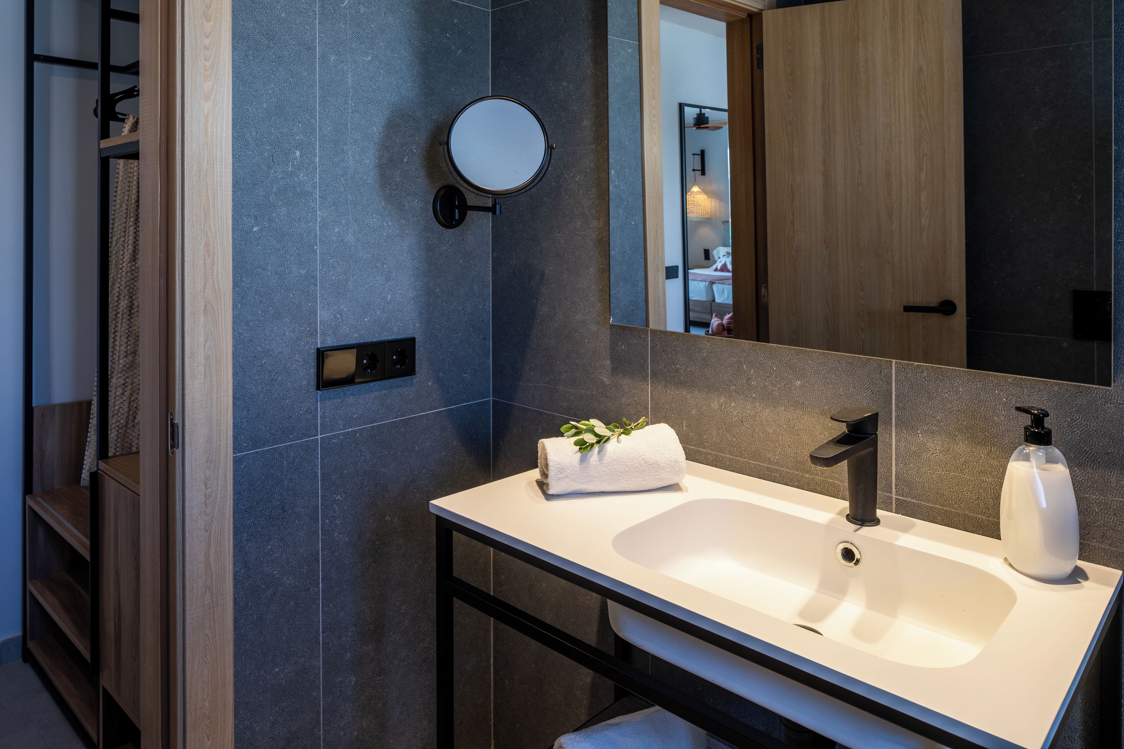 A modern bathroom with a white sink, black faucet, and a round mirror. There is a towel with a plant on the sink, and a bottle of soap on the counter.