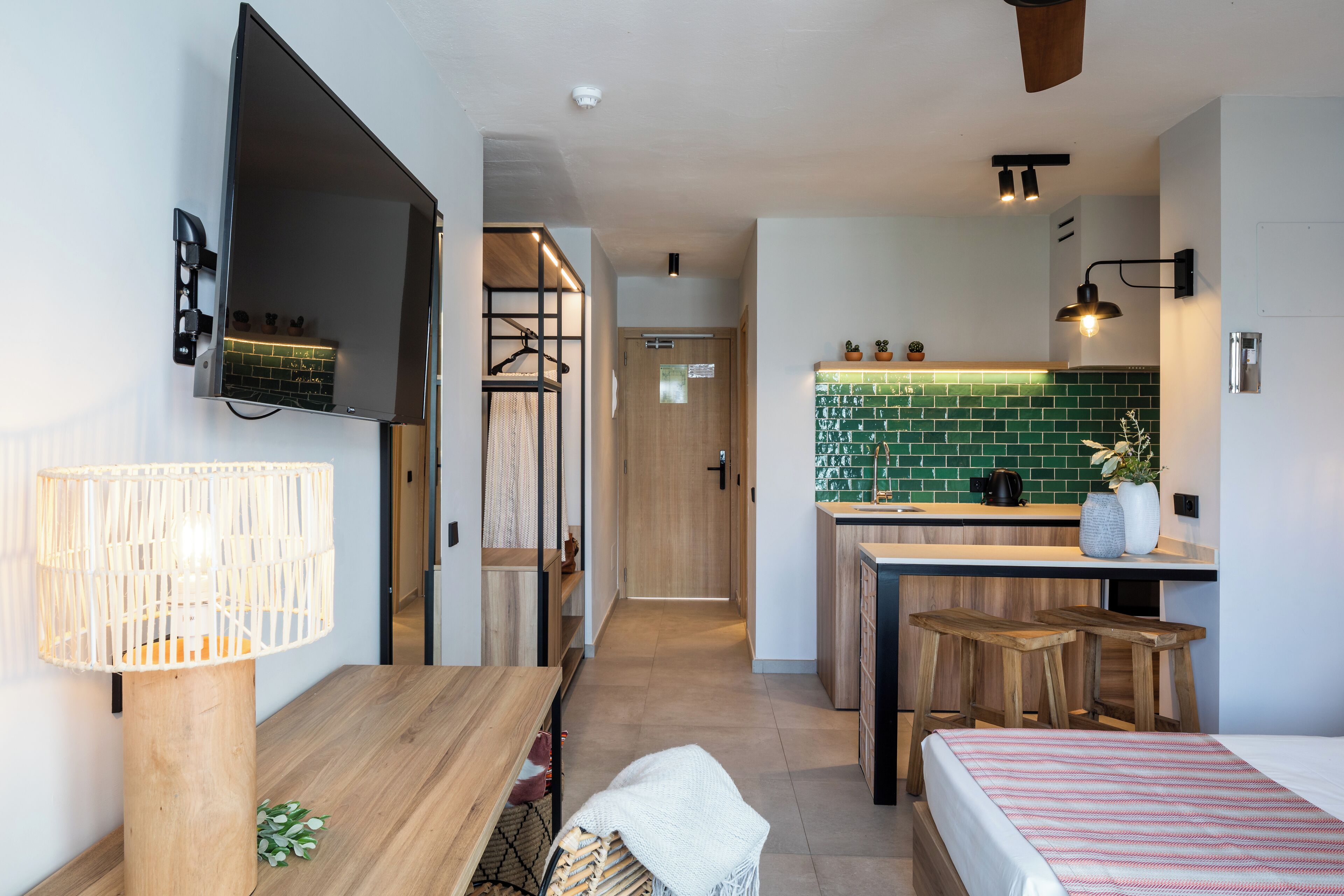 Una habitación de hotel moderna con una cama de madera, un escritorio con lámpara, un televisor montado en la pared y una kitchenette con salpicadero verde.