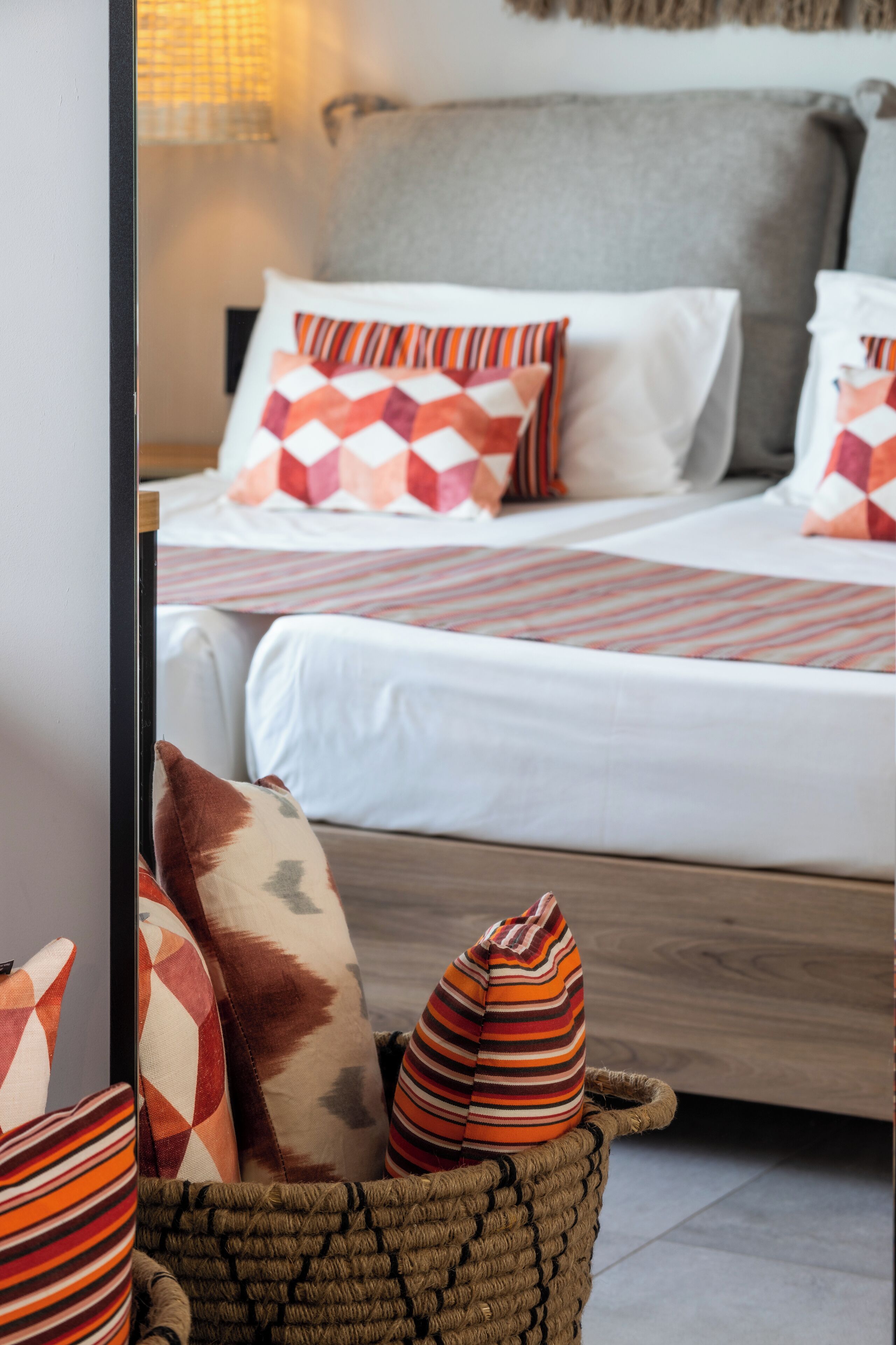 Ein ordentlich gemachtes Bett mit gemusterten Kissen, ein gewebter Korb mit zusätzlichen Kissen und eine Lampe im Hintergrund.