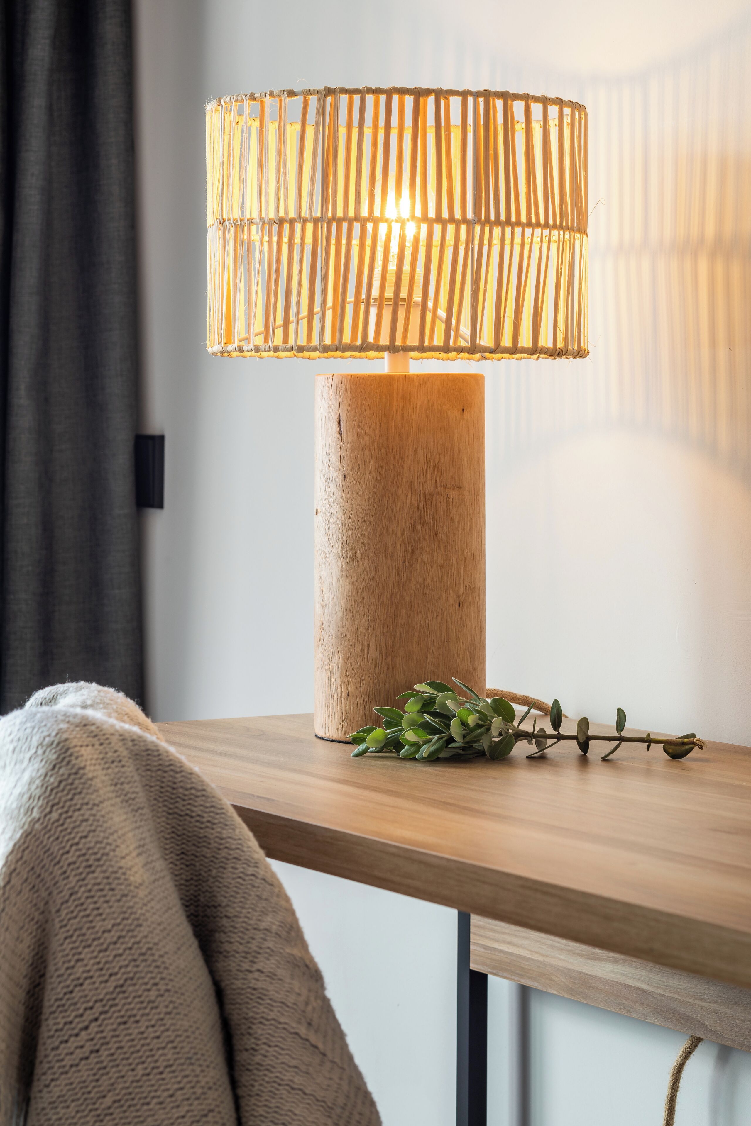 Una mesa de madera con una lámpara trenzada, una planta verde y una manta gris.