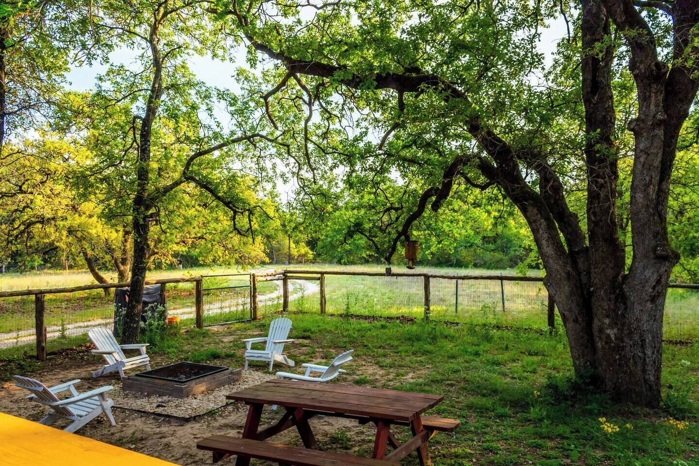 A picnic area with a wooden table and four Adirondack chairs, surrounded by a fence and trees.