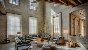 Een ruime woonkamer met een hoge plafond, zichtbare bakstenen muren en grote ramen die natuurlijk licht binnenlaten.