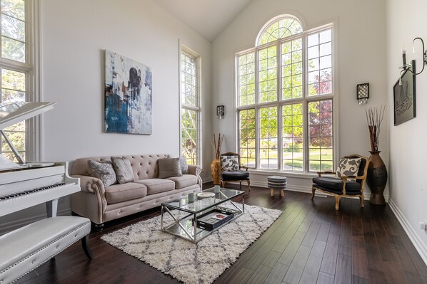 A spacious living room with a grand piano, a beige sofa, a glass coffee table, and large windows offering a view of the outdoors. - GLOBALSTAY. Unique 5BR Villa. Swimming Pool. HOT TUB (Lincoln)