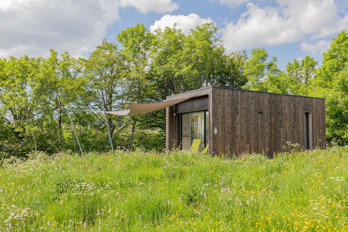 Une cabane en bois moderne, située dans un champ verdoyant, dotée d’une grande fenêtre et d’un auvent.
