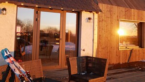 Un bâtiment en bois avec un toit en tuiles, de grandes portes vitrées et une terrasse aménagée avec des meubles en osier.