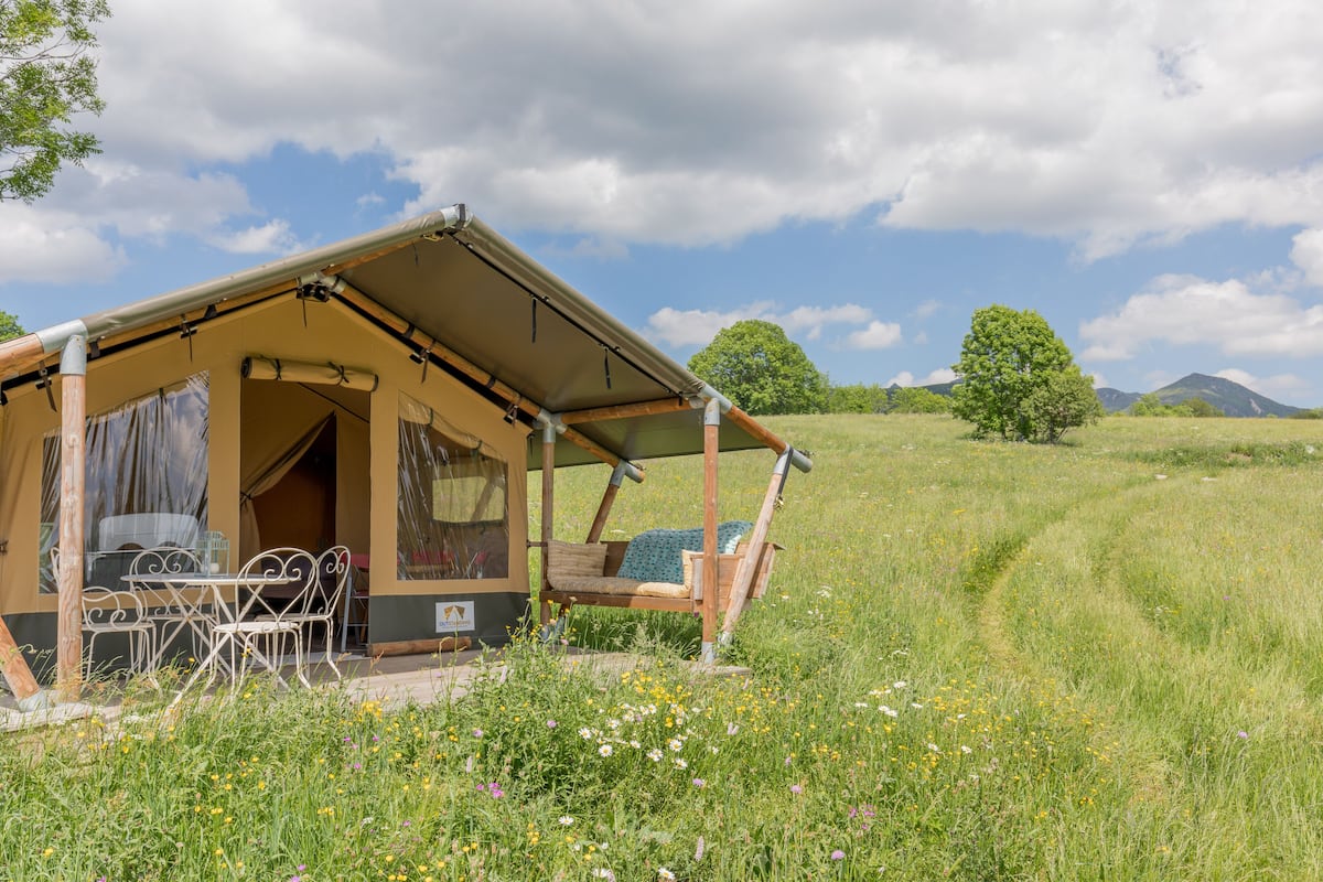 Un hébergement rustique aménagé dans une tente, comprenant un coin repas et un lit simple, situé dans un champ verdoyant avec une montagne en toile de fond.