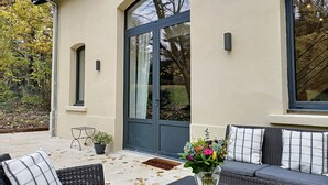 Een overdekt terras met buitenmeubels, een plant in pot en een groot raam.
