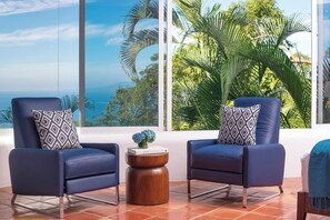 Zwei dunkelblaue Sessel mit gemusterten Kissen, ein kleiner Holztisch und Blick auf Palmen und Meer durch große Fenster.