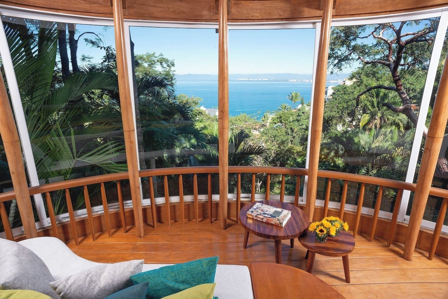 A room with a curved wooden balcony, a round wooden table with a book and flowers, and a view of the ocean and greenery.