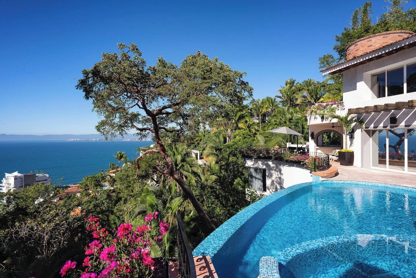 Une vue sur la piscine, avec une vue dégagée sur la mer, entourée d’une végétation luxuriante et d’un bâtiment moderne doté de grandes fenêtres.