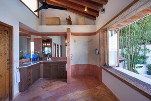 A bathroom with a large mirror, wooden cabinets, a shower area, and a window with a view of greenery.