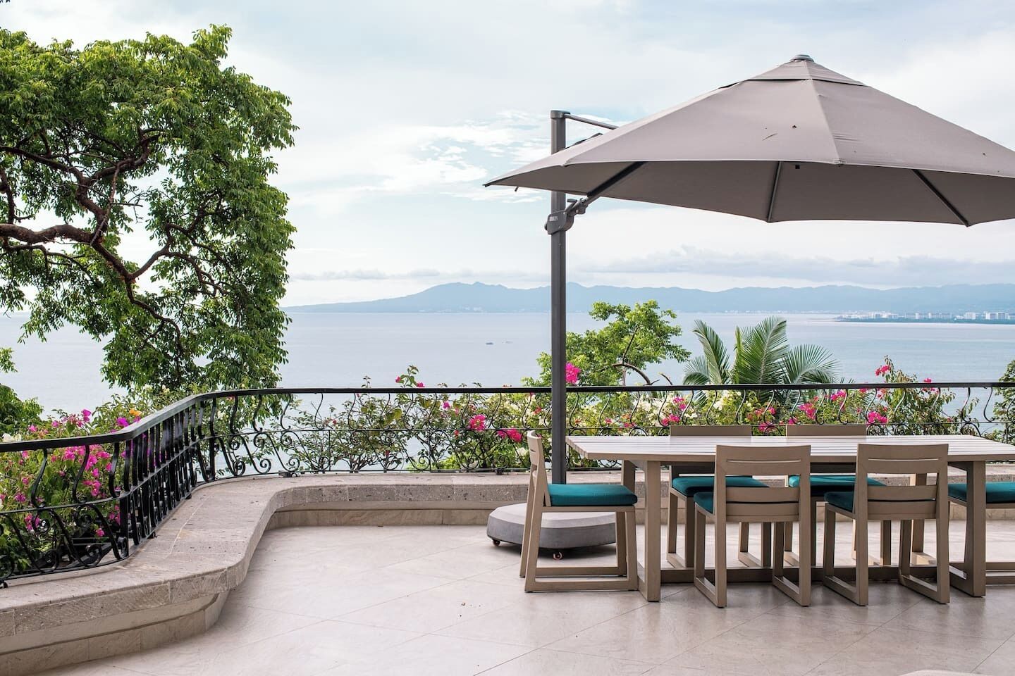 Une terrasse extérieure avec une table et des chaises, un grand parasol et une vue sur la mer et les montagnes.