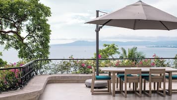Une terrasse extérieure avec une table et des chaises, un grand parasol et une vue sur la mer et les montagnes.
