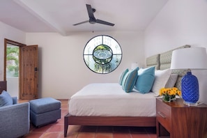 Un dormitorio con cama, ventilador de techo, ventana con diseño decorativo, mesita de noche con lámpara y una silla.