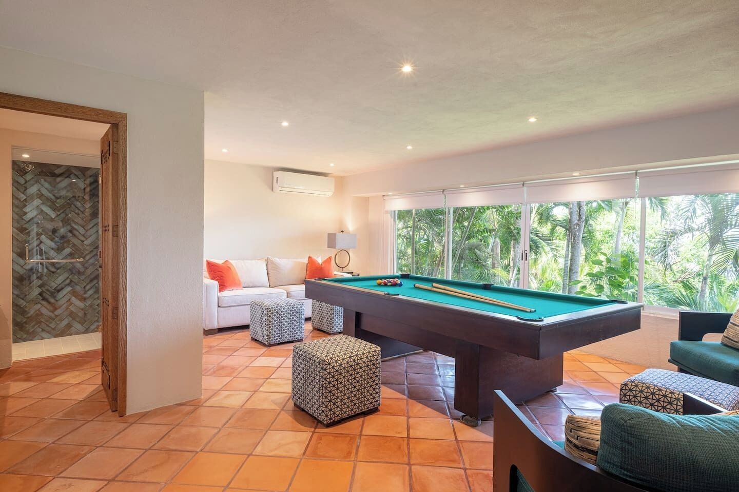 Une table de billard dans une pièce où se trouvent un canapé blanc, des poufs à motifs et de grandes fenêtres donnant sur un paysage verdoyant.