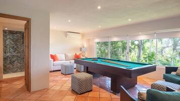 Une table de billard dans une pièce où se trouvent un canapé blanc, des poufs à motifs et de grandes fenêtres donnant sur un paysage verdoyant.