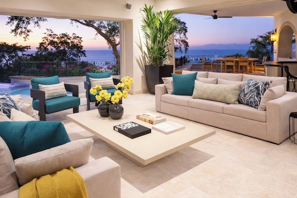A modern living room with a view of the sea, a sofa, chairs, a coffee table with books and flowers, and a dining area with a table and chairs.