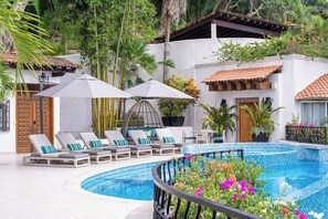 A pool area with lounge chairs and umbrellas, surrounded by greenery and a white building with a tiled roof.
