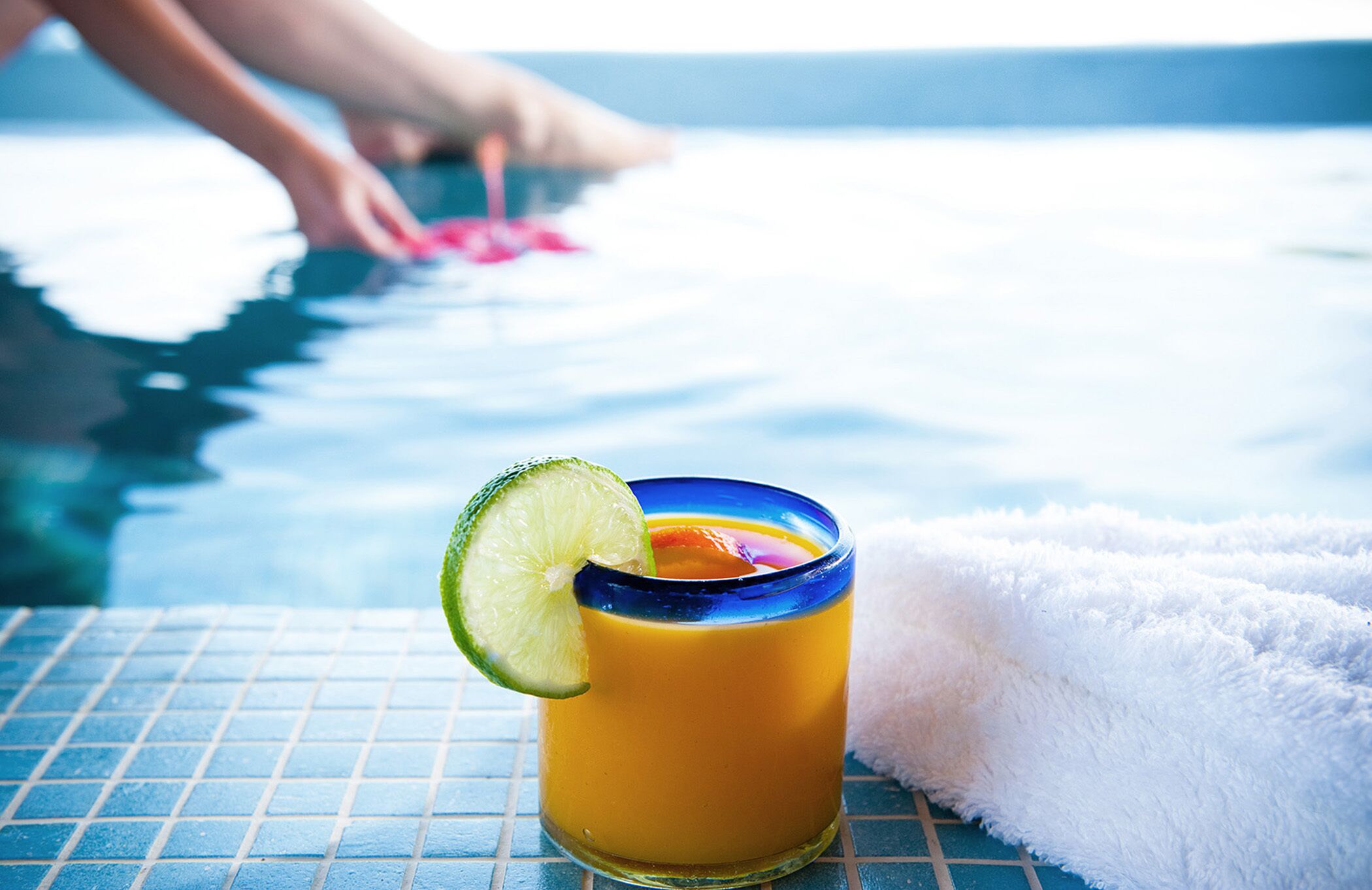 Un verre de boisson à l’orange, garni d’une tranche de citron vert, posé sur une table au bord d’une piscine carrelée.