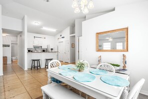Un interior luminoso y blanco con una mesa de comedor dispuesta para cuatro, una cocina al fondo y un ventilador de techo.