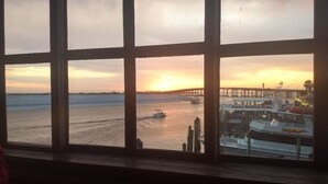 Vista pela janela com um pôr do sol sobre a água, um cais e um barco atracado em um porto.