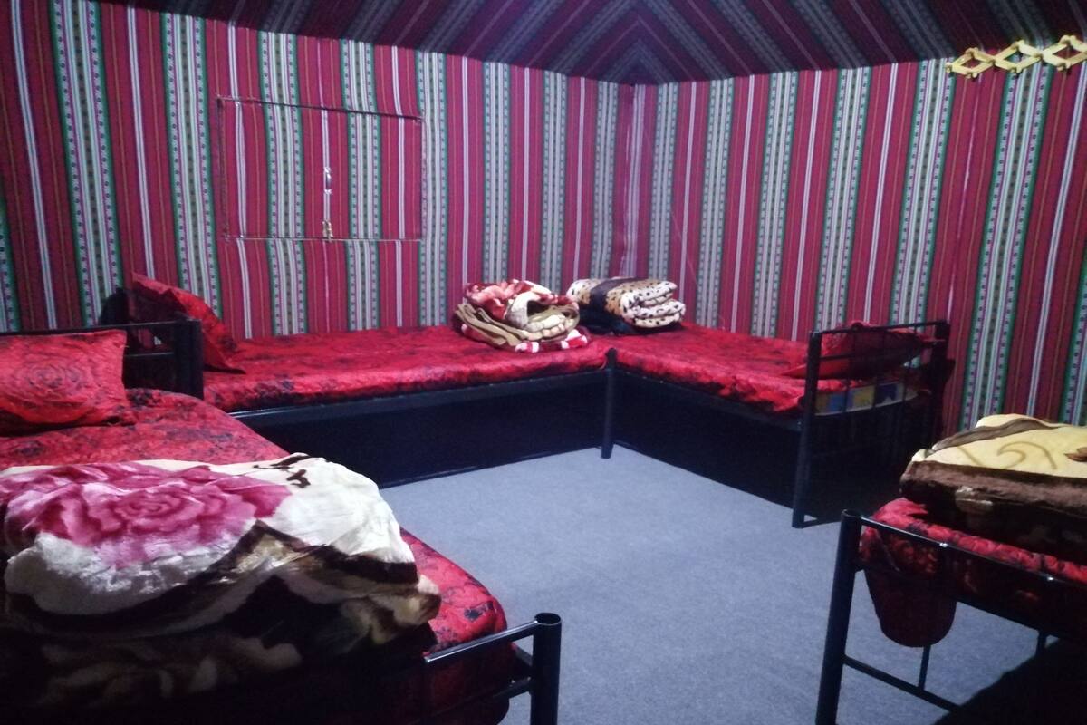 Ein Zimmer mit zwei Betten, eines mit roter Bettdecke und das andere mit weißer. Ein Sessel und ein kleiner Tisch mit gelber Tischdecke stehen bereit. Die Wände sind mit einem Streifenmuster versehen.