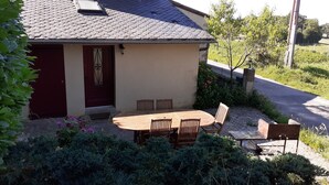 Une maison avec un toit en tuiles, une porte rouge, une table et des chaises en bois, et un jardin où poussent différentes plantes.