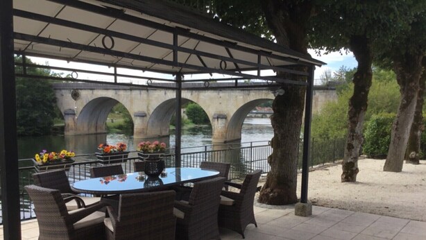 Zona salotto esterna riparata, con mobili in vimini e composizioni floreali, affacciata su un fiume e un ponte.
