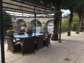 A covered outdoor seating area with wicker furniture and flower arrangements, overlooking a river and a bridge. - Petit Chateau of ‘La Belle Époque’ era on the banks of the River L’Isle. (Saint-Astier)