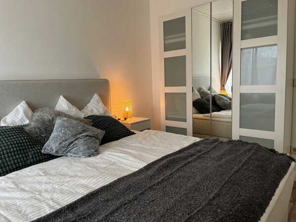 A neatly made bed with a grey headboard, white and grey pillows, and a dark grey throw blanket. A bedside lamp with a yellow shade is on the nightstand beside the bed.