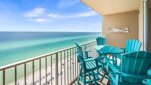 A balcony with turquoise chairs and a table, overlooking a beach with clear turquoise water and a sandy shore.