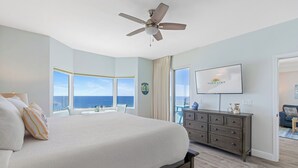 A bedroom with a large bed, a ceiling fan, a TV, and a view of the ocean.