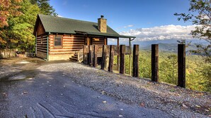 Une cabane en rondins avec une cheminée en pierre, une véranda et une allée de gravier menant à une clôture en bois, d’où l’on peut admirer un paysage montagneux.