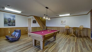 Une table de billard dans une pièce dotée d’un plancher en bois, d’un fauteuil inclinable et d’un escalier.