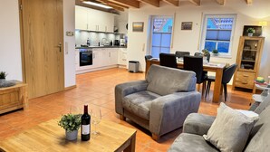 Amplia sala de estar con un sofá, una mesa de centro de madera y una mesa de comedor con sillas. Se ve una cocina con armarios blancos y una zona de comedor con ventana al fondo.