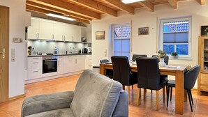 Una cocina moderna con armarios blancos, un comedor con mesa y sillas de madera, y un sofá gris en primer plano.
