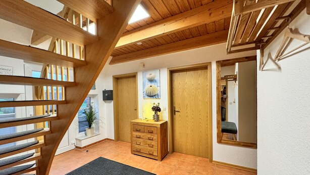 Una escalera de madera que sube a un piso superior, una puerta de madera con un pequeño ventanal, y un suelo de baldosas.