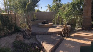 Un espace extérieur aménagé en terrasse, avec des sièges disposés en cercle autour d’un foyer et entouré de palmiers.