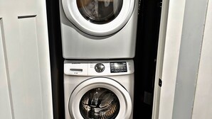 A compact laundry room with two stacked washing machines, a control panel, and a door. - Cozy Charming •Art 🎨 • Food🌮 Close to Train 🚉 • Parking! (Chicago)