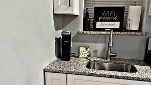 A kitchen with white cabinets, a granite countertop, a stainless steel sink, a Keurig coffee maker, a sign with a password, and a window with blinds. - Cozy Charming •Art 🎨 • Food🌮 Close to Train 🚉 • Parking! (Chicago)